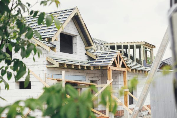 Unfinished modern farmhouse building. Wooden roof framing of mansard with dormer and vapor barrier on aerated concrete block walls with windows. Rafters and beams on blocks. New house construction